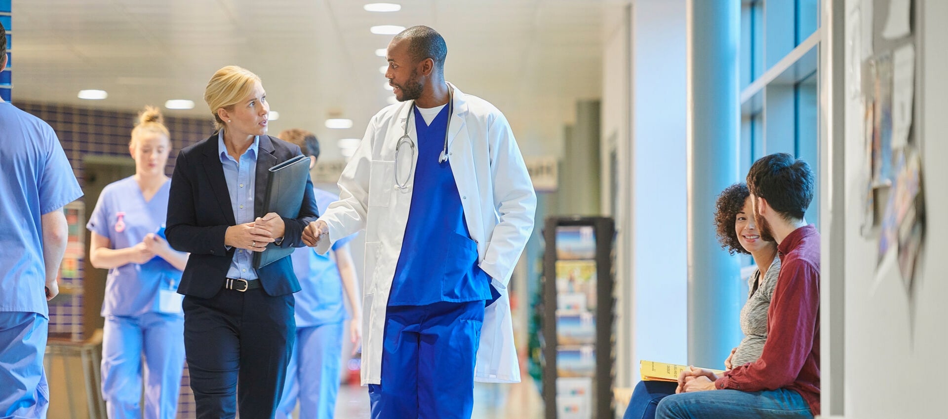 Healthcare administrator and doctor walk through hospital in conversation.
